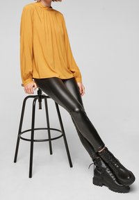 Senapsgul långärmad blus med veckdetaljer, i kombination med glänsande svarta leggings och svarta snörkängor på en barstol.