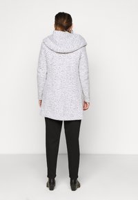 Cappotto grigio chiaro con macchie, dotato di un ampio cappuccio, maniche lunghe e un orlo leggermente svasato. Abbinato a pantaloni neri aderenti e scarpe con tacchi.