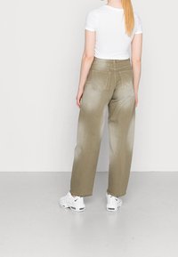 Wijduitlopende jeans in een verloopkleur olijfgroen, met twee achterzakken, versleten randjes en een verweerde textuur; gecombineerd met witte sneakers.
