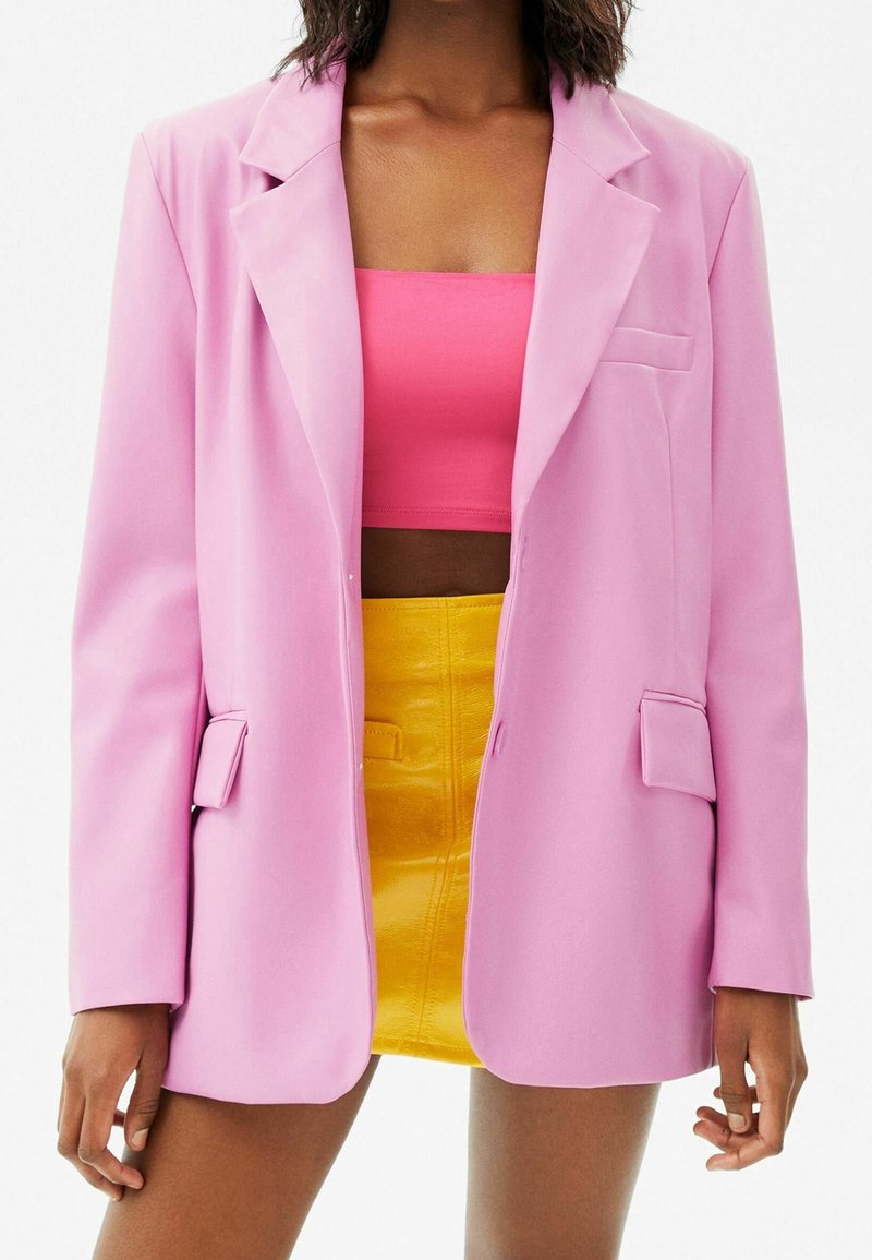 Femme portant un blazer rose pastel, un top tube rose vif et une mini-jupe jaune taille haute, vue de la mi-cuisse au cou.