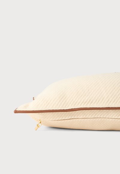 Coussin beige doux texturé avec bordure marron et fermeture éclair dorée visible sur un fond clair uni.