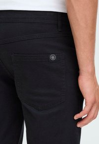Svart denimbukser med en baklomme som har en sirkulær logo-tag. Stoffet virker glatt med en lett glans, laget med en skreddersydd passform.