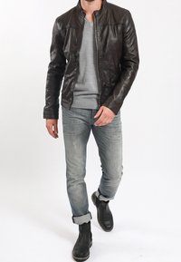 Brun läderjacka med ståkrage, kombinerad med en ljusgrå V-hals tröja och blekblå jeans med uppvikt ärm och svarta stövlar.