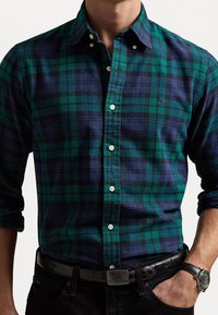 Grün- und marineblaues Karomuster-Flanellhemd mit Button-down-Kragen, langen Ärmeln, weißen Knöpfen und kleinem Logo. Getragen mit schwarzem Gürtel und Uhr.