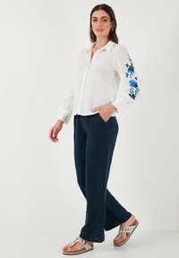 Chemise blanche en lin à boutons avec broderies florales bleues sur les manches, associée à un pantalon large bleu marine et des sandales argentées.