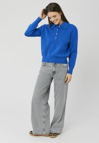 Pull en maille bleu avec un col à revers et détails boutonnés, associé à un jean large gris clair et des ballerines à imprimé animal.