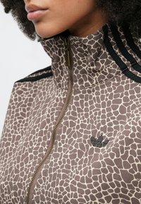 Καφέ και μπεζ ζακέτα με φερμουάρ και σχέδιο καμηλοπάρδαλης, ψηλ ός γιακάς, μαύρες ρίγες στους ώμους και μικρό μαύρο λογότυπο κοντά στο φερμουάρ.