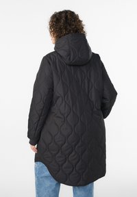 Persona ar cirtainiem matiem, valkājot garu melnu kapuci ar šūtu virsjaku un gaiši zilas džinsas, stāvot ar muguru pret baltu vienkrāsainu fonu.