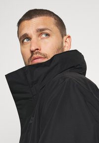 Veste imperméable noire avec un col montant. Texture lisse, design minimaliste, avec un logo près de l'épaule.