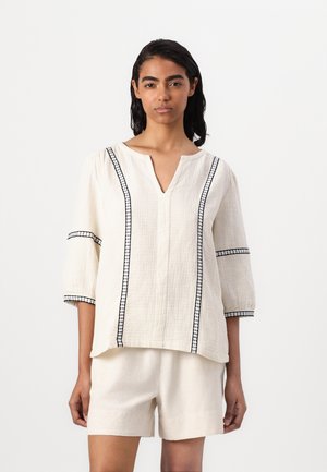 Hvid linnedbluse med V-udskæring, korte puffærmer og sorte detaljer langs sømmene og manchetterne. Sammen med beige shorts.
