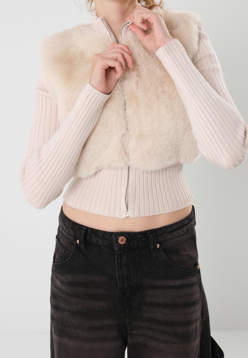 Femme fermant la fermeture éclair d'un pull côtelé beige avec gilet en fausse fourrure intégré, portant un jean noir taille haute, sur fond neutre.