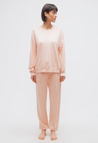 Γυναίκα που στέκεται ξυπόλητη, φοράει άνετο, ανοιχτό ροδακινί μακρυμάνικο τοπ και αντίστοιχο παντελόνι, κοιτάζοντας προς τα αριστερά της, μπροστά σε απλό λευκό φόντο.