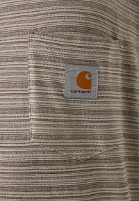 Striped fabric shirt featuring horizontal brown and cream stripes, a patch pocket, and a Carhartt logo tag in orange and gray.