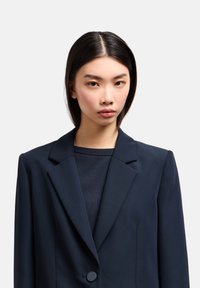 Blazer blu navy con rever a lancia, chiusura a un bottone e spalle strutturate. Tessuto liscio con una vestibilità su misura.