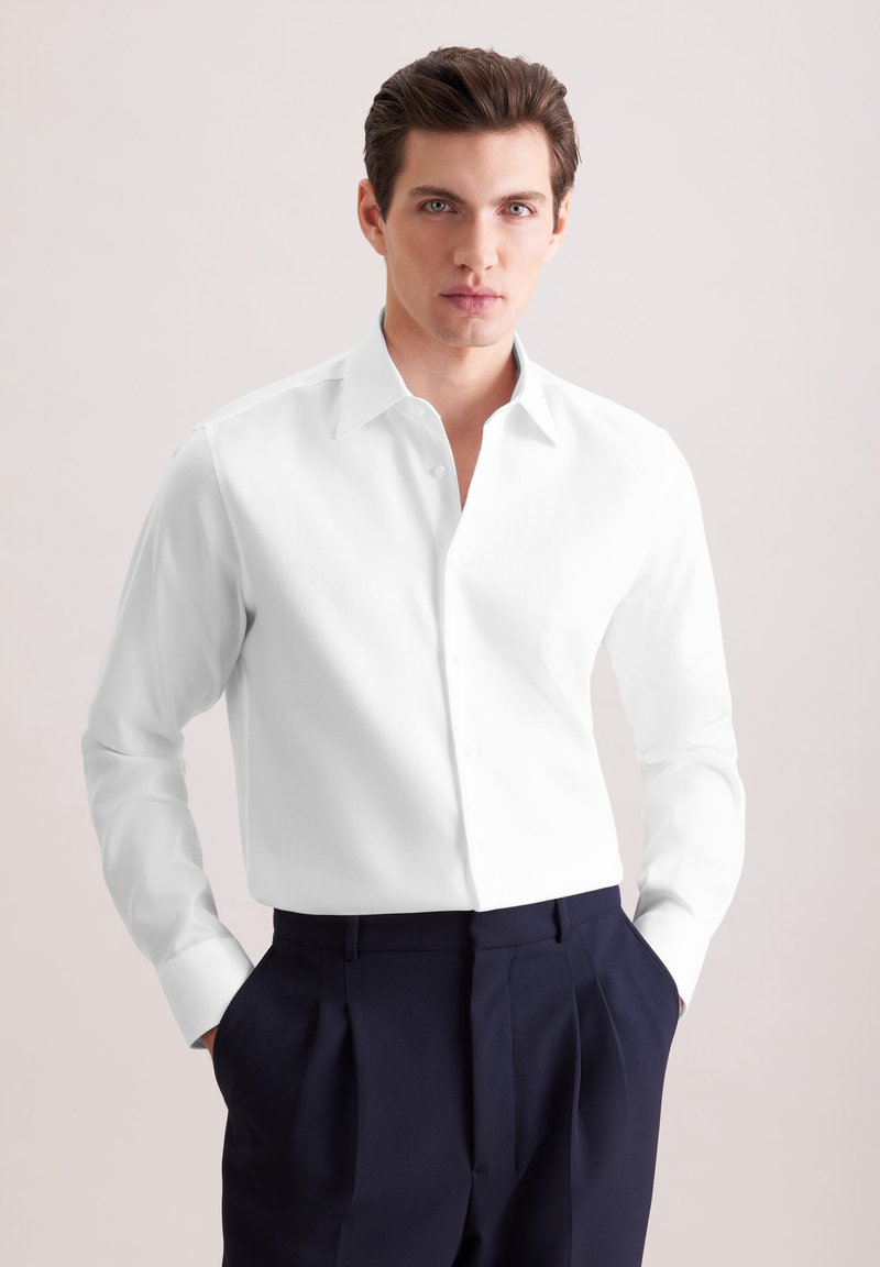 Jeune homme portant une chemise blanche à manches longues boutonnée et un pantalon foncé plissé, debout les mains dans les poches devant un fond neutre.