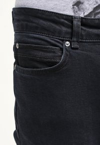 Dark denim jeans with a close-up view showing the waistband, coin pocket, and stitching details. The texture appears smooth and uniform.