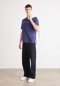 Tricou din bumbac purpuriu cu text alb, asortat cu pantaloni din denim închis și adidași albi. O hanorac de culoare deschisă este aruncat pe o umăr.