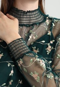 Blouse en tissu transparent vert brodé avec un col à volants et des poignets à motif quadrillé. Motifs floraux en accents rose et vert.