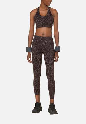 Vrouw die een donker sportbeha met patroon en legging draagt, met zwarte sportschoenen en polsgewichten, staand met het gezicht naar voren.