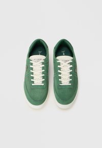 Par športnih čevljev v zeleni barvi z belimi vezalkami, belimi podplati in teksturiranim kremnim jezikom, označenim z "Lacoste Tennis."