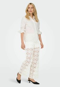 Camicia bianca con bottoni e maniche arrotolate abbinata a pantaloni bianchi in pizzo con motivo a diamante. Tacchi neri con punta a punta.