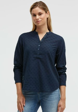 Young woman wearing a navy blue textured long-sleeve henley shirt and light blue jeans, standing against a plain background.