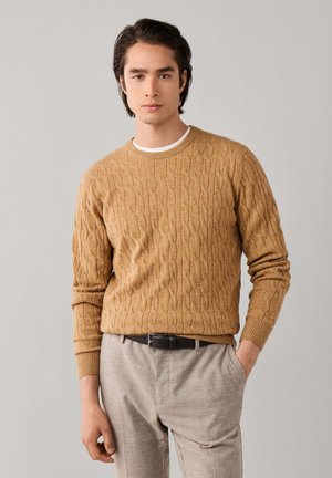 Jonge man met donker haar die een beige kabelgebreide trui, lichte geruite broek en een zwarte riem draagt, staand met één hand in de zak.