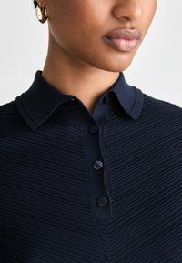 Polo navy con strisce testurizzate, colletto classico e patta con tre bottoni. I dettagli includono un tessuto morbido e bottoni abbinati.