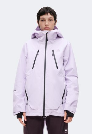 Personne portant une veste imperméable à capuche violet clair avec des fermetures éclair noires et un pantalon foncé, debout face à un fond uni.