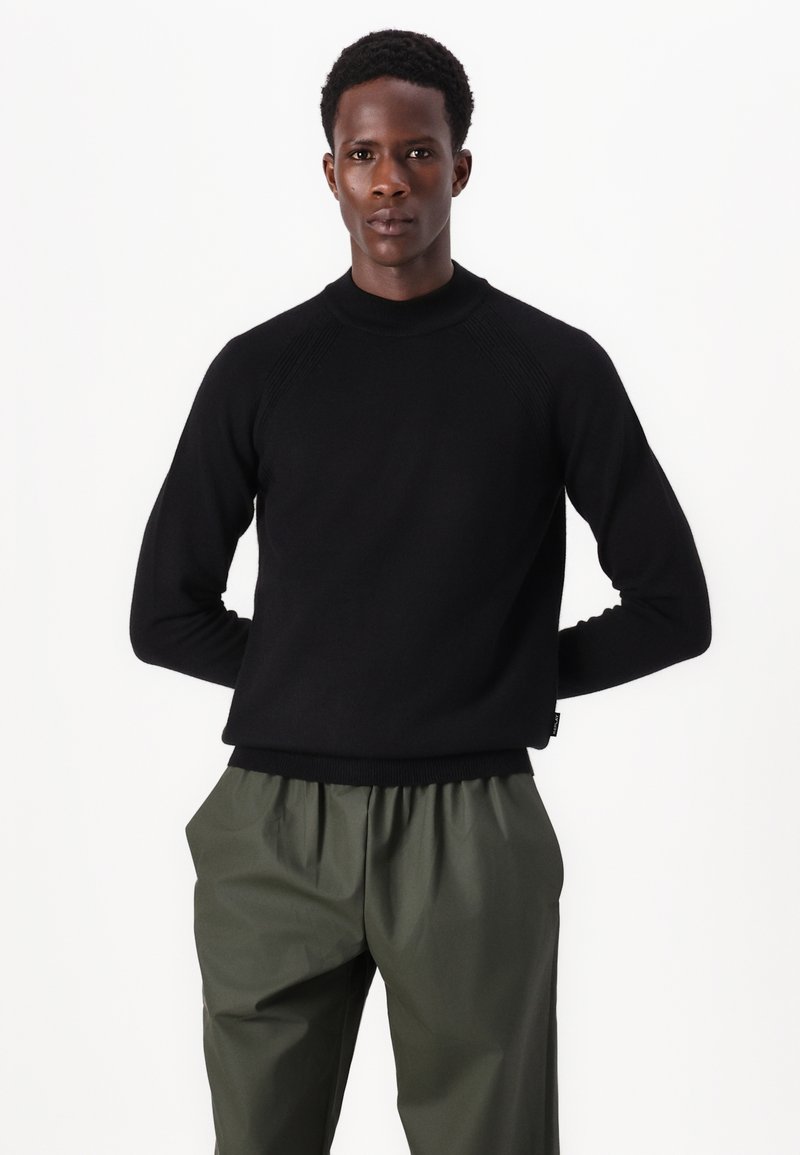 Schwarzer Rundhals-Pullover mit gerippten Details an den Schultern, kombiniert mit lockeren olivgrünen Hosen. Weicher Stoff, lässige Passform, minimalistisches Design.