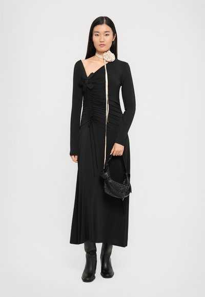 Fekete, hosszú ujjú ruha aszimmetrikus dekoltázzsal és ráncos textúrával. Virágos díszítéssel rendelkezik, és egy tüskés válltáskával van párosítva.