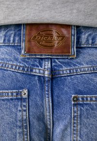 Plave traperice s smešnom kožnom etiketom u kojoj je urezano "Dickies." Sadrže dvostruko šivanje i metalne dugmadi na džepovima.