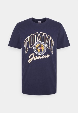 T-shirt en coton bleu marine avec un graphique "TOMMY Jeans" blanc et jaune, shorts noirs, chemise à carreaux nouée autour de la taille, baskets crème et jaune.