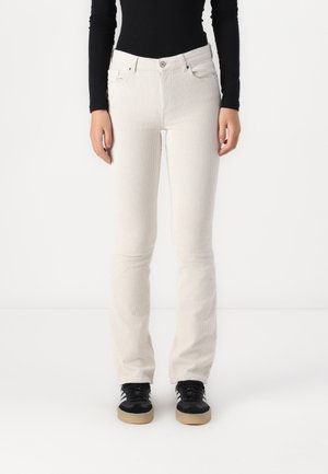 Person iført tætsiddende hvide kordbuksere, sort langærmet top og sorte sneakers med gummisåler, stående mod en hvid baggrund.