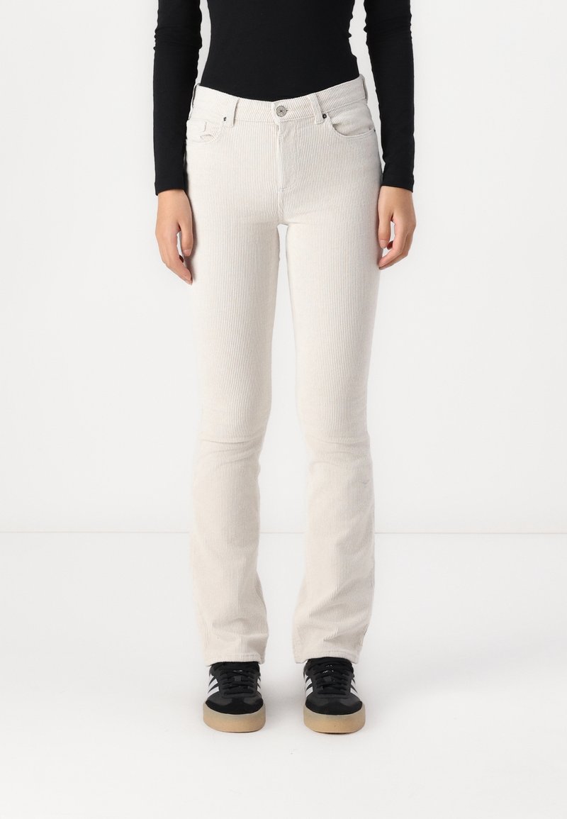 Personne portant un pantalon en velours côtelé blanc ajusté, un haut à manches longues noir et des baskets noires aux semelles en gomme, debout devant un fond blanc.