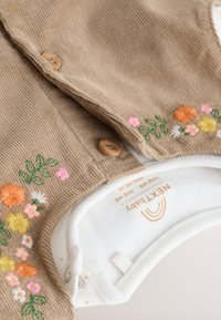 Beige corduroy baby outfit with wooden buttons and colorful floral embroidery on sleeves, paired with a white garment inside.
