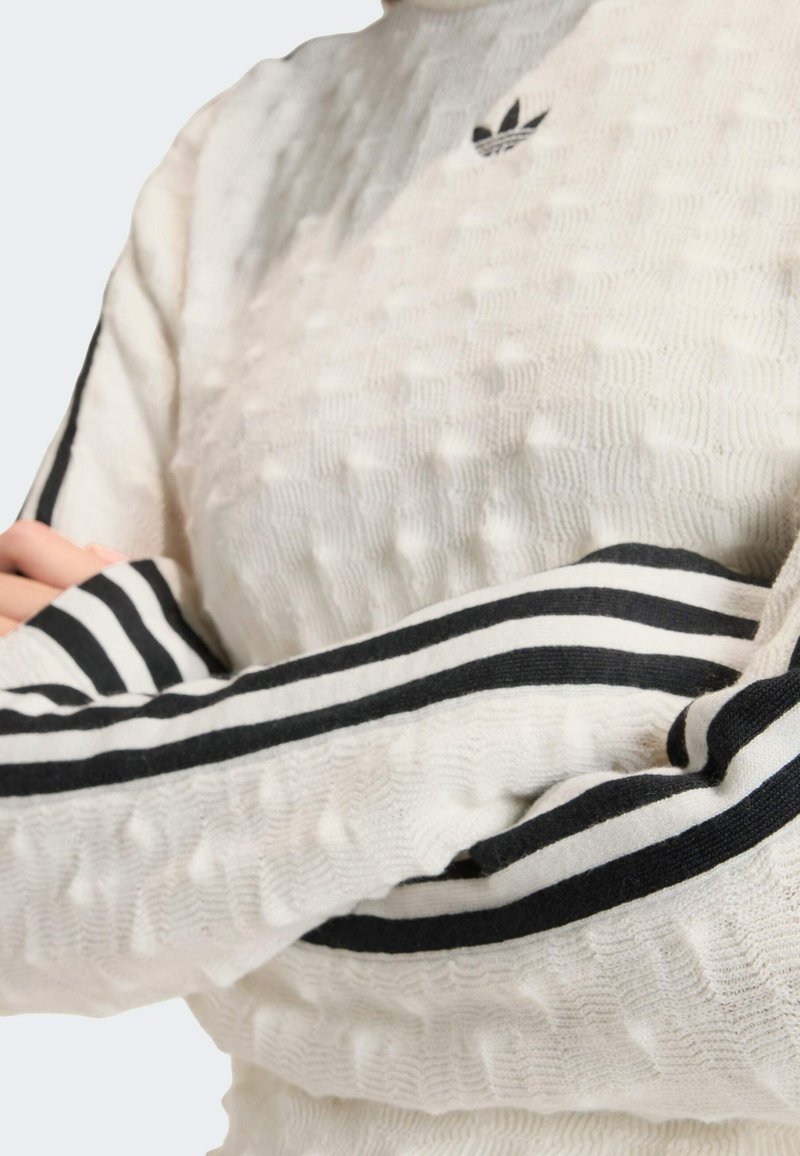 Cremefarbener, strukturierter Pullover mit schwarzen Streifen an den Ärmeln und einem gestickten Logo auf der Brust. Der Stoff wirkt weich und dick.