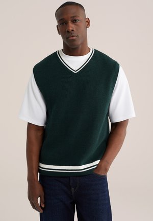 Joven con chaleco sin mangas de punto color verde oscuro sobre una camisa blanca de manga corta, con vaqueros azul oscuro, de pie de manera relajada.