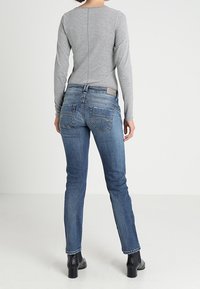 Person som bär en grå långärmad tröja, blå denimjeans med bakfickor och svarta ankelskor, står vänd bort från kameran mot en enhetlig bakgrund.