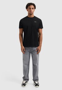 Zwarte katoenen t-shirt met korte mouwen en een ronde hals, met een klein, gouden logo op de borst. Gepaard met lichtgrijze jeans.