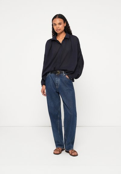 Blusa a maniche lunghe di un blu scuro, con colletto, vestibilità comoda e maniche strutturate, abbinata a jeans in denim larghi e sandali piatti.