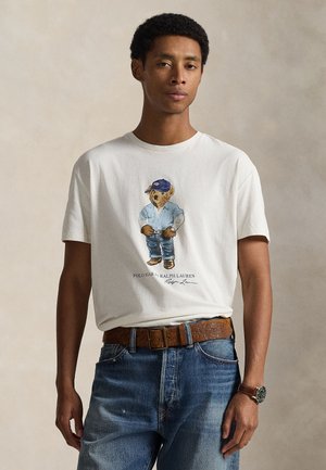 Maglietta in cotone color crema chiaro con grafica di un orso in abbigliamento casual, abbinata a jeans di denim blu e cintura marrone. Design semplice.