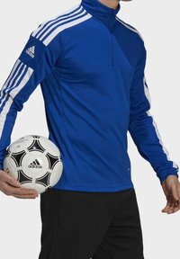 Veste de sport bleu avec des rayures blanches, col montant, fermeture éclair et manches longues, accompagnée d'un ballon de football noir et blanc.