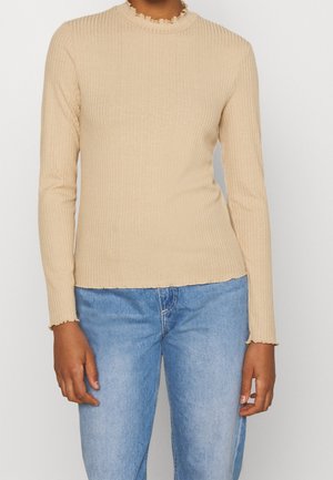 Beżowa prążkowana bluzka z długim rękawem, z falbaną przy okrągłym dekolcie i prostym dołem. W zestawie z jasnoniebieskimi dżinsami. Gładka tekstura.