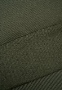 Ciemnozielona dziana tkanina o prążkowanej teksturze i gładkich akcentach. Charakteryzuje się różnorodnymi wzorami, w tym odcinkiem o delikatniejszym splocie i szerszym pasem.