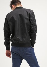 Mężczyzna w czarnej kurtce bombowcu i niebieskich dżinsach, odwrócony plecami, z jedną ręką w luźnej pozycji i lekko patrzący w dół.