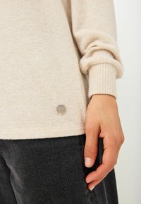 Beige stickad tröja med ribbade muddar, med en liten silverfärgad metallaccent på fållen. Bärs med mörkgrå jeans.