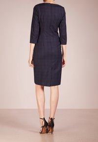 Femme se tenant de dos, portant une robe ajustée noire marine arrivant aux genoux et des sandales à talons noirs avec des brides de cheville.