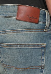 Vaqueros de mezclilla azul claro con una etiqueta de cuero marrón con texto en relieve "Pepe Jeans London." Bolsillos traseros estándar con costuras naranjas.