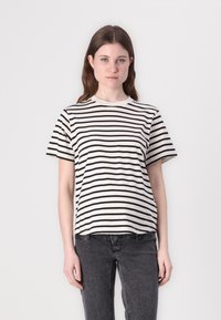 Tricou rayé în crem și negru, cu mâneci scurte, guler rotund, din material de bumbac. Modelul îl poartă cu blugi gri închis.